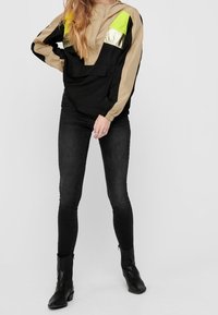 Schwarz-beige Windjacke mit Kapuze; mit neongrünen und metallischen Akzenten. Kombiniert mit schwarzen engen Jeans und Stiefeletten.