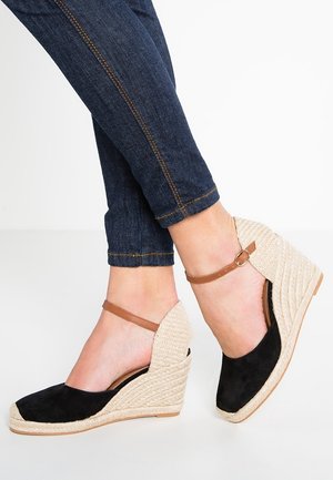 Czarne sandały na klinie zamszowe z beżowym, plecionym obcasem z juty, regulowanym brązowym paskiem wokół kostki i odkrytym obcasem.