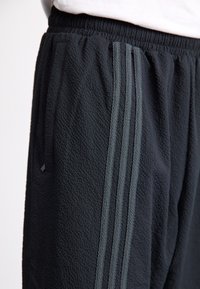 Svarta joggingbyxor i texturerat tyg med ribbad midjeresår och tre vertikala grå ränder längs sidan. Inklusive sidofickor.