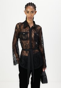 Camicia nera in pizzo con maniche lunghe, colletto a punta, due tasche sul petto e una cintura annodata in vita; design floreale trasparente, texture liscia.