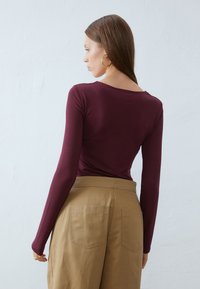 Burgundy long-sleeve top with a scooped neckline, paired with beige high-waisted pants featuring two back pockets. Smooth, fitted fabric.