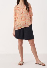 Blouse en voile fleurie et pêche avec un décolleté en V et des manches retroussées, associée à des shorts navy et des sandales à nœud de couleur claire.