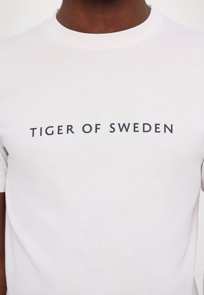 Weißes Baumwoll-T-Shirt mit kurzen Ärmeln und Rundhalsausschnitt, mit einem dunkelblauen Druck "TIGER OF SWEDEN" auf der Vorderseite.