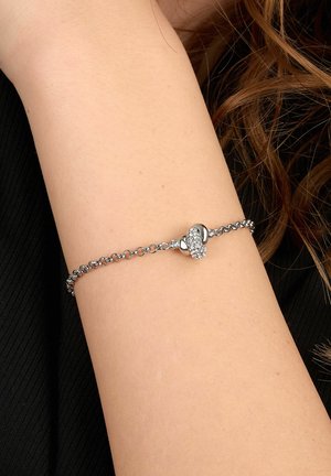 Bracciale con catena d'argento e ciondolo a forma di cuore adornato con piccole pietre scintillanti indossato sul polso di una persona.