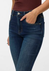 Jeans de mezclilla azul oscuro con cintura de altura media, con bolsillos delanteros, costuras naranjas en contraste y cierre con botón metálico.