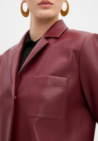Blazer in eco-pelle bordeaux con collarino a scollo a V, taschino singolo sul petto e texture liscia, abbinato a un top nero sottostante.