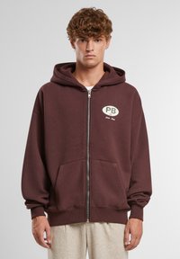 Burgundská mikina na zips s kapucňou, predným logom, vreckami na kapse, a rebrovanými manžetami a lemom. Vyrobená z mäkkého materiálu s hladkou textúrou.