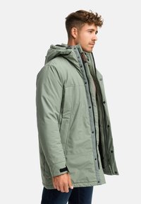 Giacca verde imbottita con cappuccio, zip anteriore e bottoni a pressione. Presenta polsini elastici e un design trapuntato. Materiale leggero.
