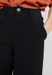 Pantalons noirs ajustés avec une texture lisse, une devant plissé, des poches latérales et une fermeture à bouton avec un bouton contrastant.