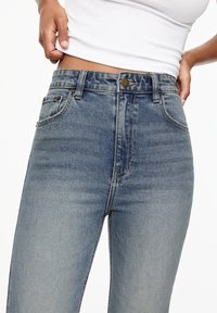 Jean bleu taille haute en denim, avec un effet délavé, cinq poches et un bouton métallique à la taille.