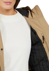 Beige Jacke mit einem schwarzen strukturierten Innenfutter, offen, um einen weißen gerippten Sweater darunter zu zeigen. Die Jacke hat einen hohen Kragen und eine mit Reißverschluss ausgestattete Tasche.