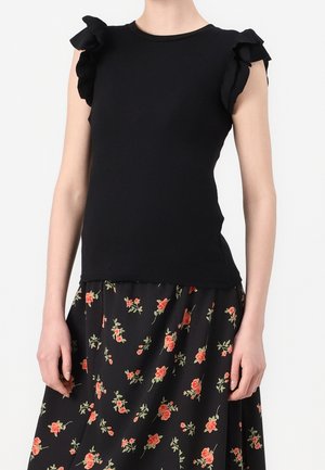 Kobieta w czarnej, bezrękawnikowej bluzce z falbanami na ramionach oraz czarnej spódnicy z małym czerwonym wzorem kwiatowym na jednolitym białym tle.