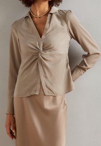Femme en blouse en satin beige avec un détail noué à l'avant et une jupe assortie, portant un collier en chaîne dorée, se tenant devant un mur uni.