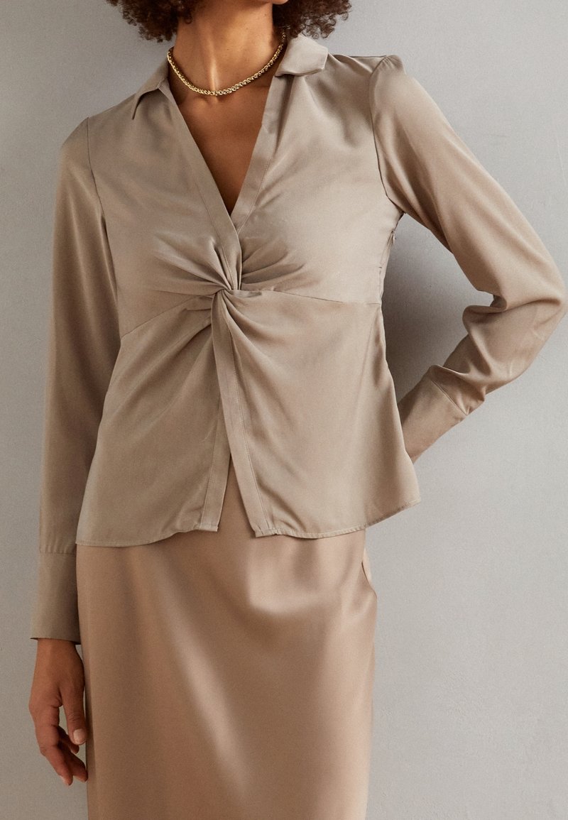 Femme en blouse en satin beige avec un détail noué à l'avant et une jupe assortie, portant un collier en chaîne dorée, se tenant devant un mur uni.