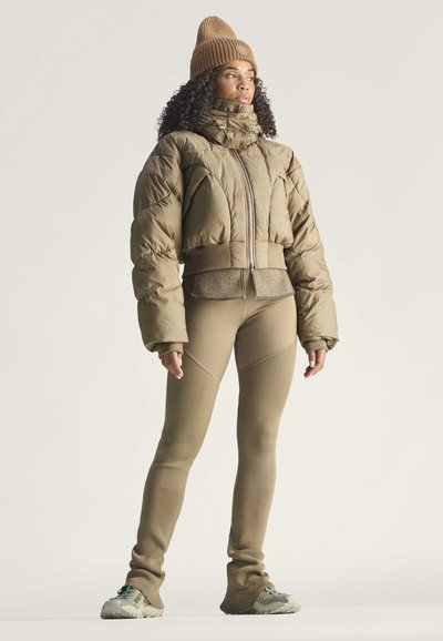 Mujer de pie que lleva un gorro marrón, una chaqueta acolchada oliva, leggings beige ajustados y zapatillas grises sobre un fondo liso.