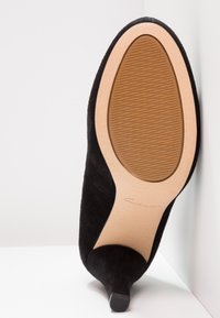 Chaussure à talon haut en suède noir avec un bout pointu et une semelle en caoutchouc texturé. La semelle est beige et présente un détail de marque discret.