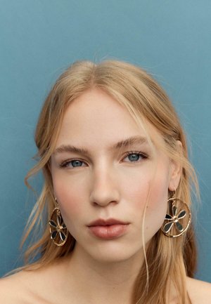 Femme blonde aux yeux bleus portant de grandes boucles d'oreilles en forme de fleur dorée sur un fond bleu uni.