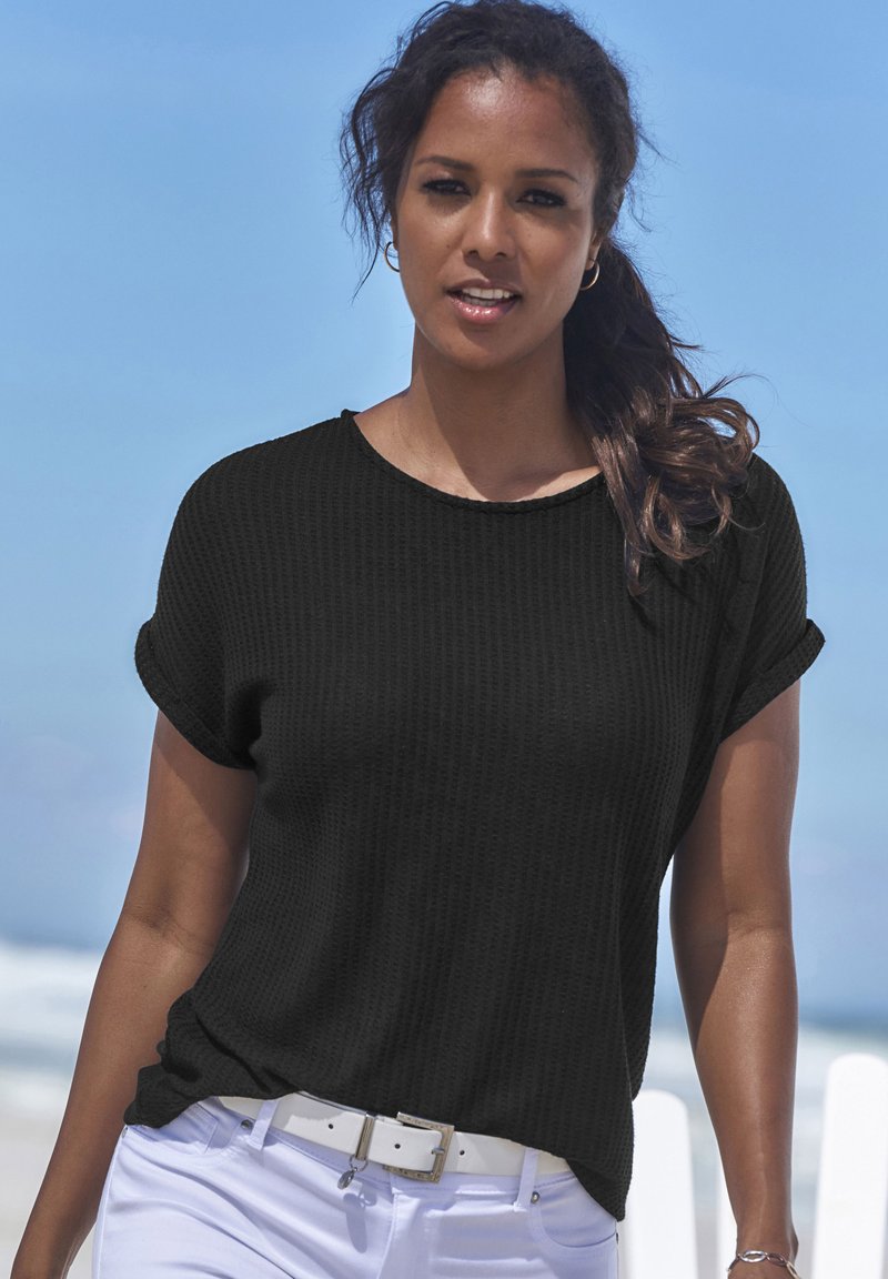 Mustaline, lühikeste varrukatega, tekstuuriga pullover koos ümmarguse kaelusega, kantud valgete pükste ja valge vöödiga. Väljas taust, sinine taevas.