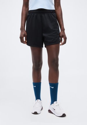 Osoba oblečená v čiernych športových šortkách, bielych teniskách a modrých Nike ponožkách stojaca oproti jednoduchému pozadiu.