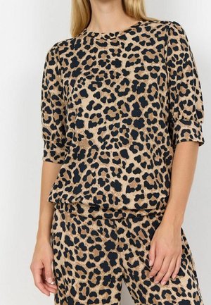 Femme portant un haut imprimé léopard beige et noir avec des manches jusqu'au coude et un pantalon assorti, debout devant un fond blanc uni.