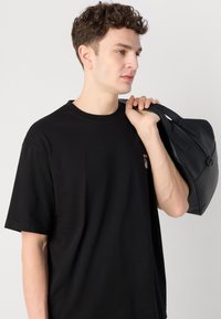 Černá kožená taška s dvojitými uchy, zaobleným tvarem, hladkým povrchem a minimalistickým designem. Nosená přes rameno osobou v černém tričku.