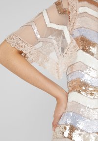 Robe à paillettes présentant un motif en chevrons dans des tons de rose poudré, bronze et bleu clair. Manches en dentelle transparente avec des détails délicats sur les bords.