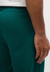 Türkiz színű pamut melegítőnadrág sima textúrával, rugalmas derékbőséggel és testhezálló fazonnal, egy kéz a combon pihen.