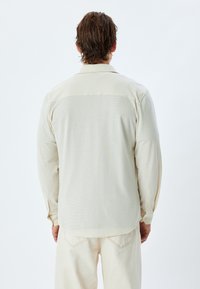 Camisa de manga larga beige claro con un patrón texturizado, puños abotonados y un panel superior liso de contraste. Vista trasera mostrada.
