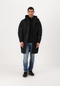 Svart lang parka-kåpe med glidelås og trykknappe-lukking, med hette og to frontlommer, båret over en mørk genser og jeans.