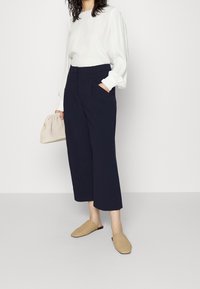 Mujer vistiendo una blusa blanca de manga larga, pantalones cortos anchos de pierna navy, zapatos de lona beige de tipo slip-on, y sosteniendo un bolso de mano tejido beige.