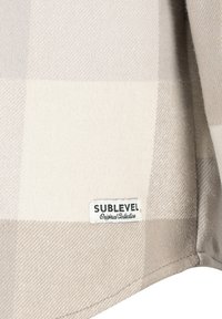 Beige kariertes Hemd aus weichem, strukturiertem Stoff. Mit einem Etikett „SUBLEVEL Original Collection“ nahe dem Saum.