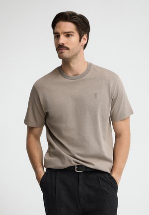 Uomo con capelli scuri e baffi, che indossa una t-shirt taupe a maniche corte e pantaloni neri con le mani nelle tasche, su uno sfondo uniforme.
