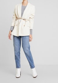 Blazer doppiopetto color crema con vita cintata, abbinato a jeans azzurri chiari e stivaletti bianchi lucidi alla caviglia. La camicia a righe spunta dai polsini.
