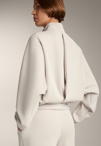 Vaaleanharmaa ribattupaita, jossa on korkea kaulus, laskeutuvat olkapäät ja takasaumassa laskos, yhdistettynä vastaaviin ribattuihin housuihin ja rentoon istuvuuteen.