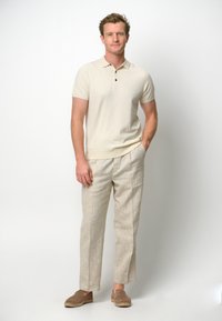 Beige pikétröja med treknappars knäppning, i kombination med ljusa linbyxor. Bruna slip-on-skor fulländar outfiten.