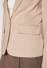 Blazer beige texturé avec une coupe ajustée, fermeture à un bouton et deux poches avant. Tissu lisse, revers structurés, et longueur raccourcie.
