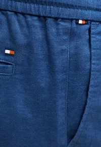 Κοντινή λήψη μπλε παντελονιού από ύφασμα με πλαϊνή τσέπη και μικρές ορθογώνιες ετικέτες λογότυπου σε κόκκινο, λευκό και μαύρο κοντά στην τσέπη και τη μέση.