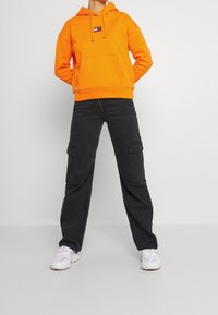 Sudadera naranja con capucha y bolsillo delantero con parche de logo, combinada con pantalones cargo negros con bolsillos laterales y corte holgado. Zapatillas blancas.