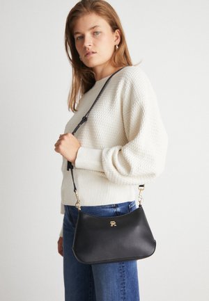 Femme portant un pull en tricot crème et un jean bleu, tenant un sac à bandoulière noir avec des accessoires dorés sur fond blanc.