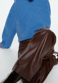 Eine Person trägt einen blauen gerippten Pullover, braune Lederhosen und schwarze Stiefel und sitzt mit einem angezogenen Knie vor einem schlichten Hintergrund.