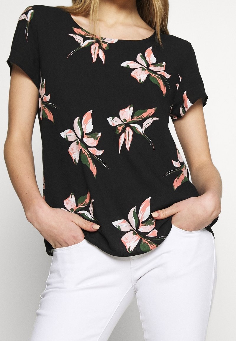 Femme portant un chemisier noir à manches courtes avec des imprimés floraux roses et verts, associé à un pantalon blanc, mains posées décontractées dans les poches.