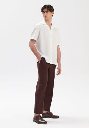 Camisa blanca de botones de manga corta, pantalones marrones ajustados y zapatos marrones oscuros. La camisa tiene un cuello relajado y una tela suave.
