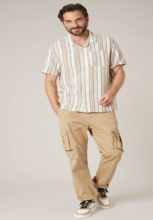 Man staat en draagt beige cargobroek, wit en beige gestreept T-shirt met korte mouwen, en witte sneakers met zwarte veters.
