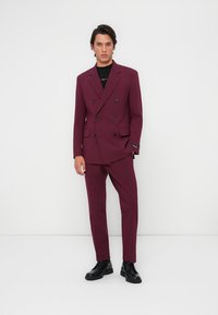 Blazer bordeaux doppiopetto con pantaloni coordinati, design aderente, texture liscia e dettagli neri. Completato con scarpe nere.