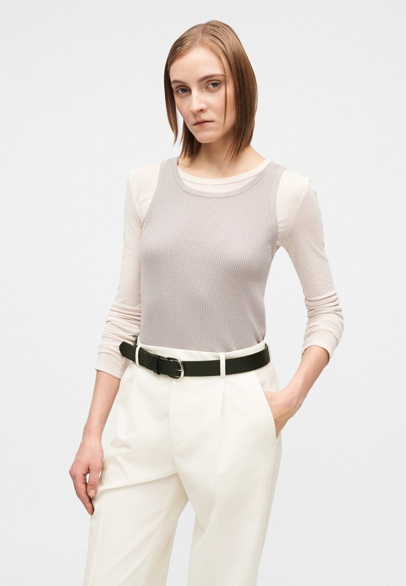 Débardeur gris côtelé superposé à un chemisier beige à manches longues, associé à un pantalon blanc taille haute et une ceinture noire avec une boucle argentée.