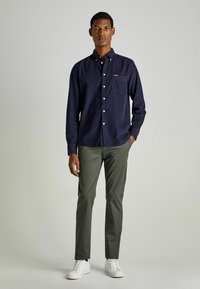 Camisa azul marino de botones con un bolsillo, combinada con pantalones verde oliva y zapatillas blancas. La camisa tiene un cuello clásico y es de manga larga.