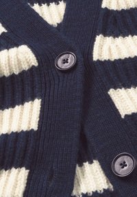 Strickjacke mit dunkelblauen und cremefarbenen Streifen, strukturiertem Stoff und einem großen schwarzen Knopf mit vier Löchern, der das Design unterstreicht.