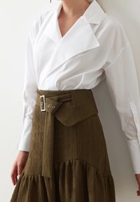 Camisa blanca de cuello con mangas largas combinada con una falda texturizada verde oliva que cuenta con un cinturón ancho y detalles de dobladillo en capas.