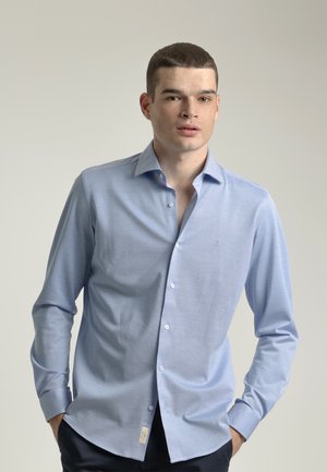 Camisa de manga larga azul claro con cuello abotonado, textura sutil y tapeta frontal. Presenta un pequeño logotipo en el pecho y en los puños.