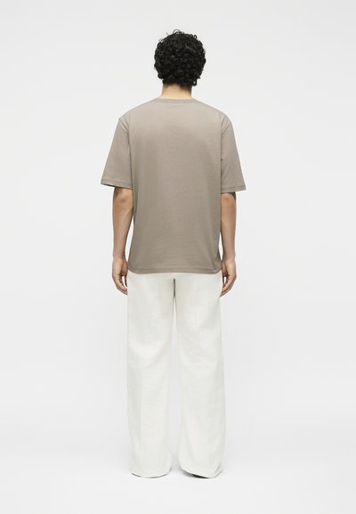 Άνδρας με σγουρά μαλλιά στέκεται με την πλάτη προς τα εμπρός φορώντας μια ταουπέ κοντομάνικη μπλούζα και άνετο λευκό παντελόνι σε απλό φόντο.